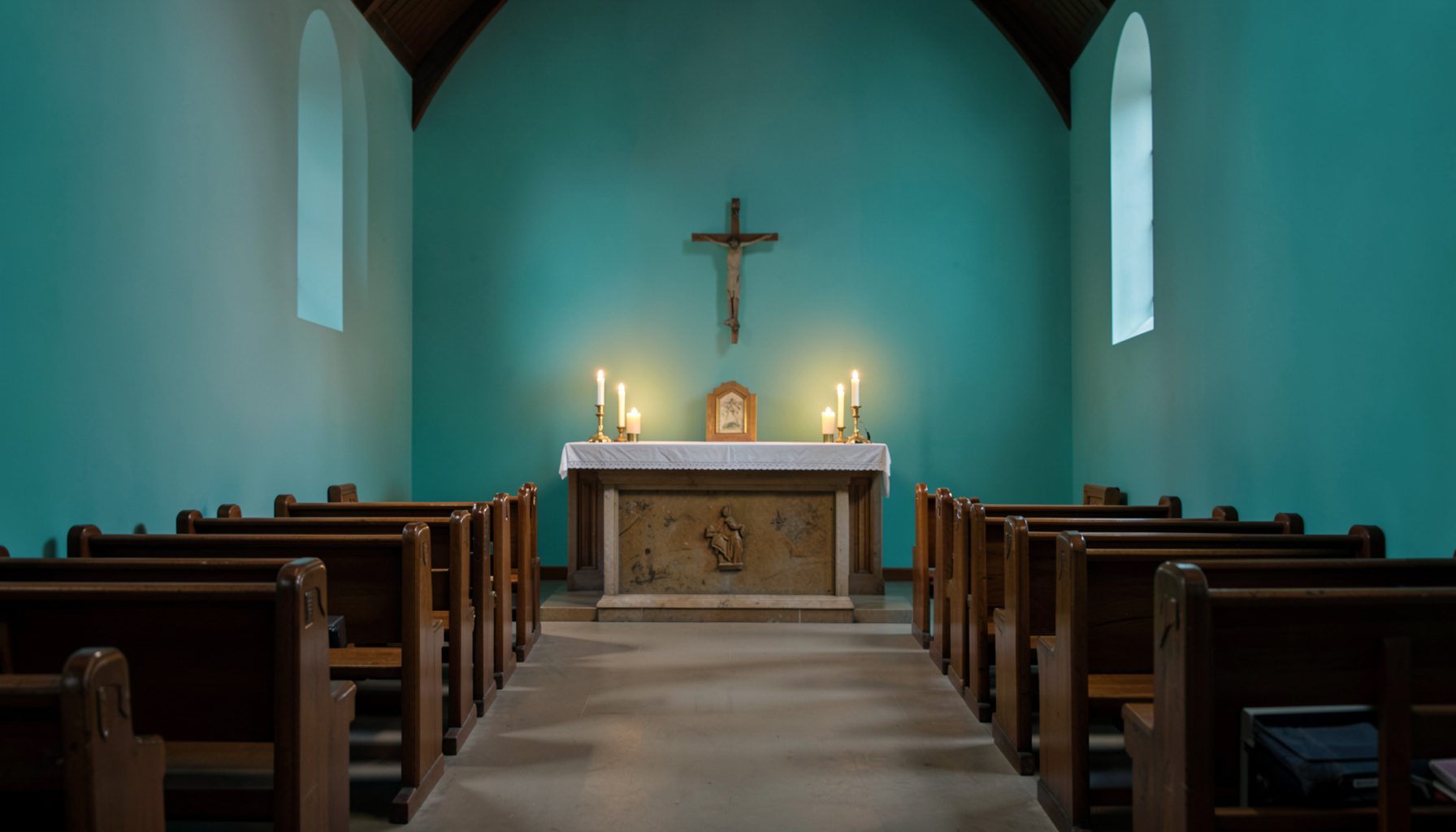 Softly lit photograph of a serene chapel interior with wooden pews, a simple altar, and warm candlelight, conveying quiet devotion and community prayer.